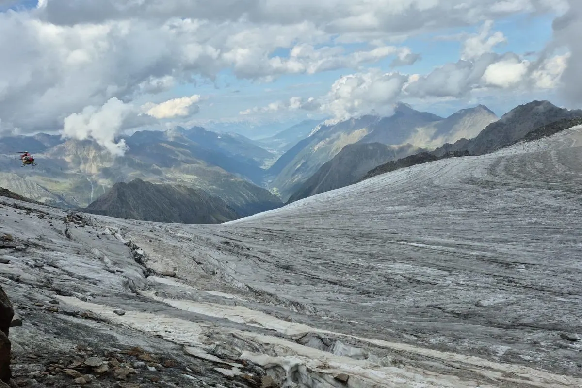 Ein roter Rettungshubschrauber fliegt über den weißen Gletscher in einer alpinen Berglandschaft mit Wolken am Himmel.