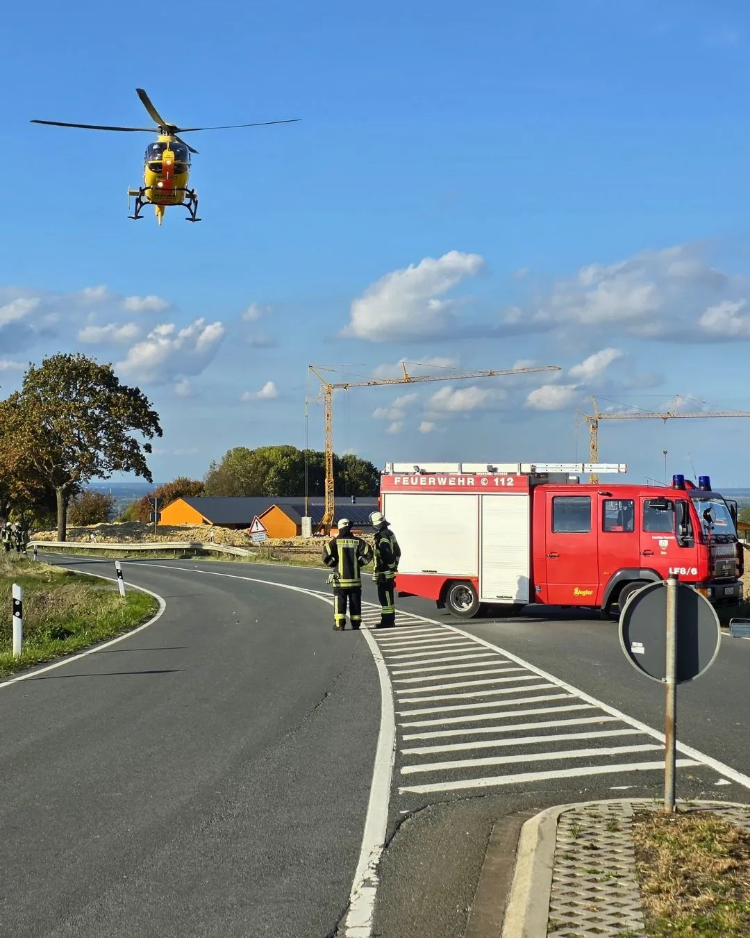 Ein gelber Rettungshubschrauber schwebt über einer kurvigen Landstraße. Im Vordergrund stehen Feuerwehrleute neben einem roten Feuerwehrauto, das die Straße blockiert.
