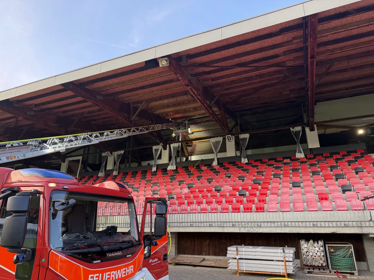 Ein rotes Feuerwehrfahrzeug mit einer ausgefahrenen Leiter steht vor den leeren Sitzreihen eines Stadions, das Dach und Holzstreben sind im Hintergrund sichtbar.