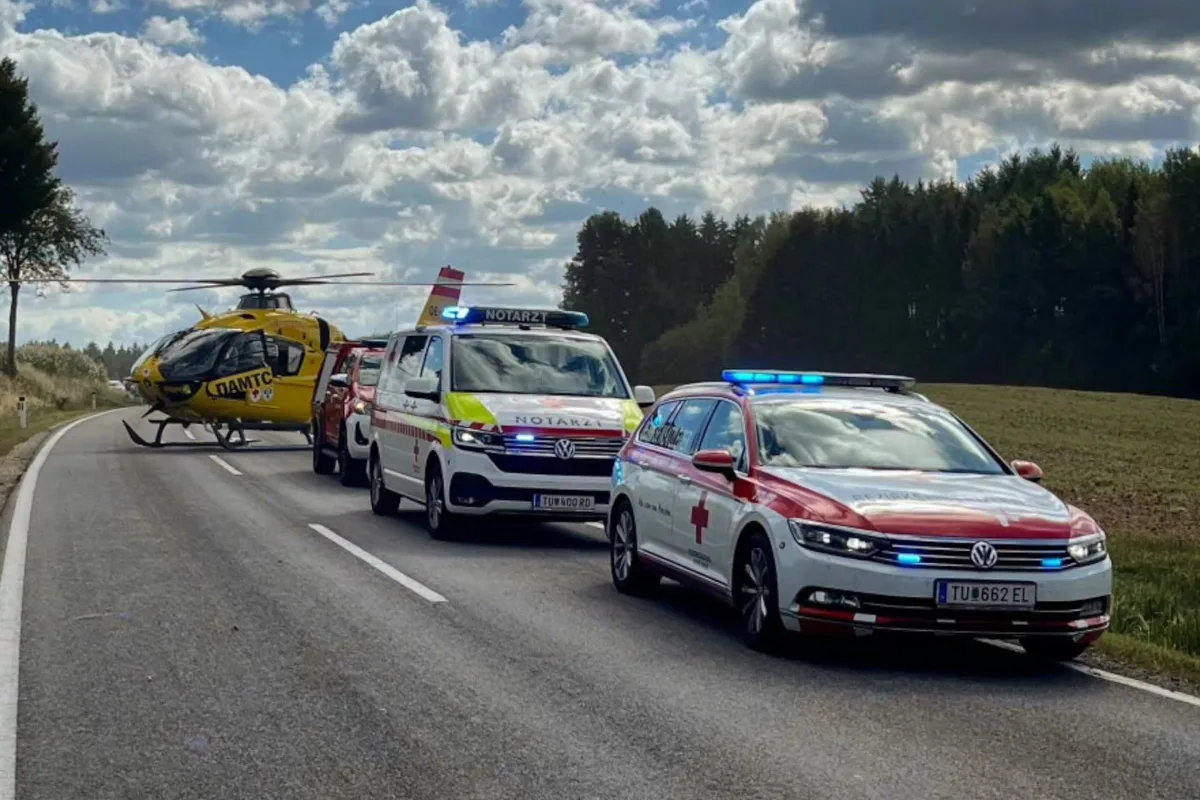 Ein gelber Rettungshubschrauber steht auf einer Landstraße, begleitet von mehreren Notfallfahrzeugen, die bereitstehen.