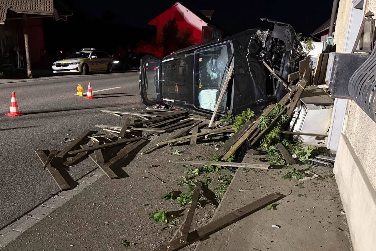 Ein schwarzes Auto liegt auf der Seite neben Trümmern auf einer Straße. Im Hintergrund sind Verkehrsleitkegel und ein weiteres Auto sichtbar.
