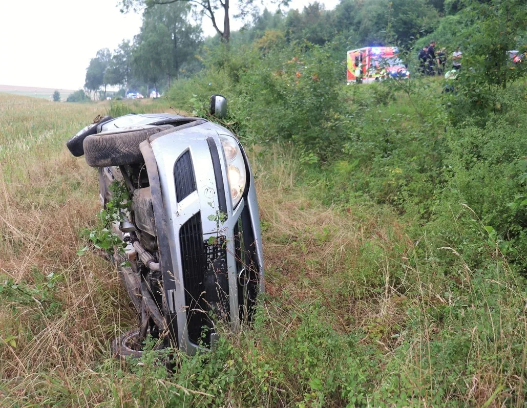 Ein silberner Mercedes liegt auf der Seite im Gebüsch, im Hintergrund sind Rettungskräfte erkennbar.