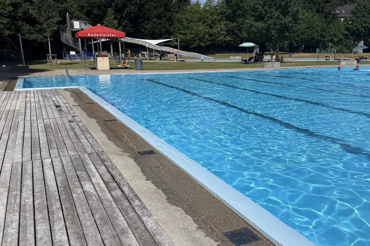 Ein großes, leeres Schwimmbecken unter freiem Himmel mit einer Wasserrutsche und einem roten Sonnenschirm im Hintergrund. Es ist ein sonniger Tag.