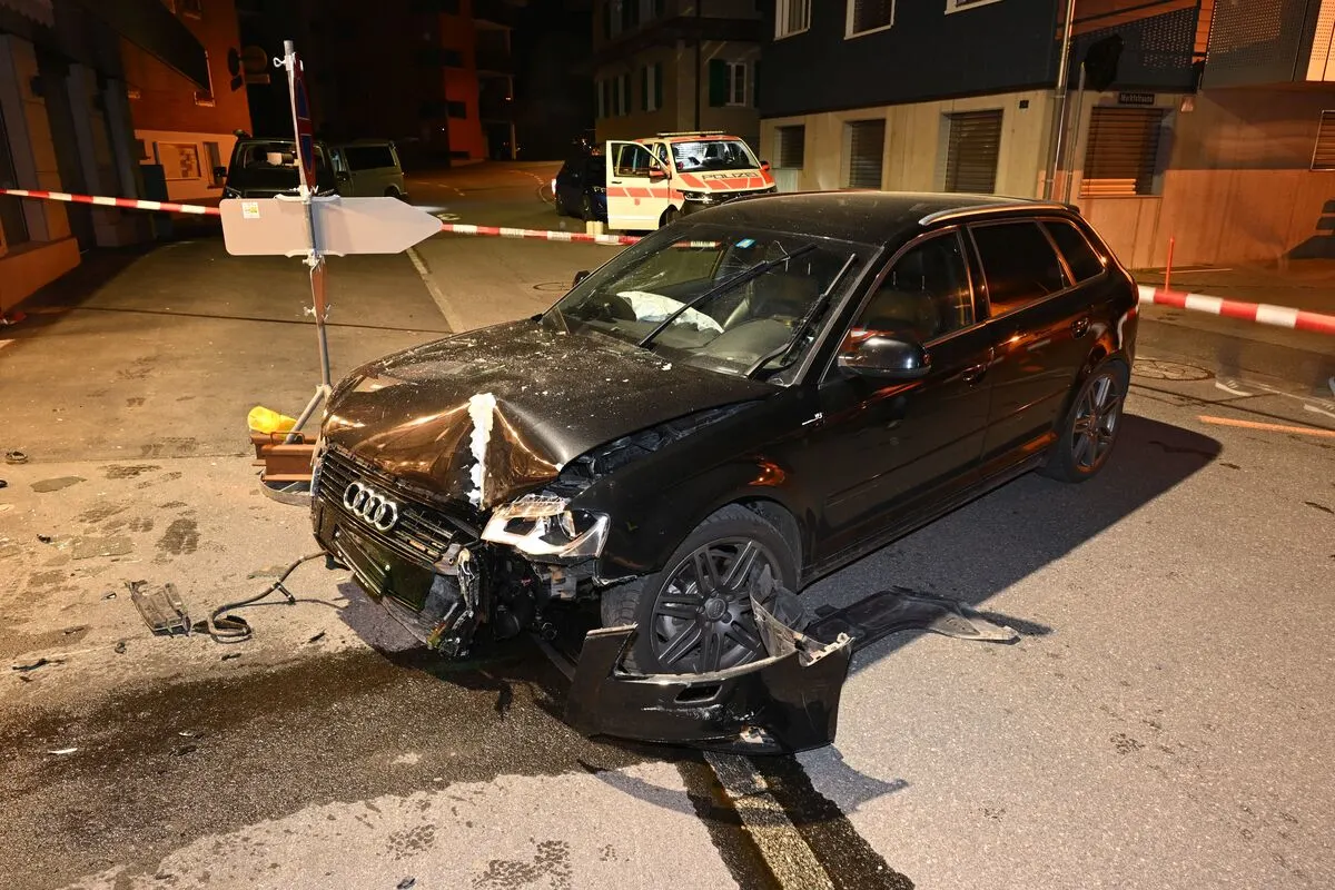Ein schwarzes Auto mit erheblichem Frontschaden steht auf einer Straße. Im Hintergrund ist ein Polizeiabsperrband zu sehen, sowie Feuerwehr- oder Polizeifahrzeuge.