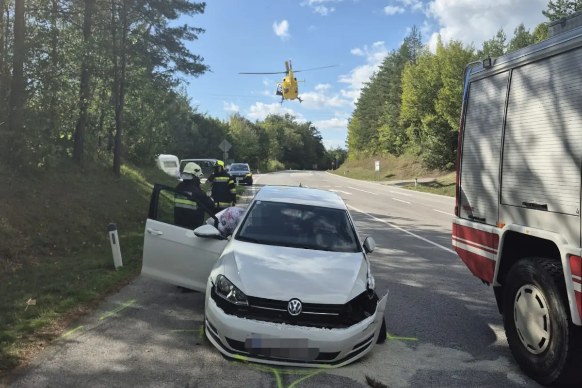 Ein weißer VW mit beschädigter Front steht auf einer Landstraße, daneben Feuerwehrkräfte und ein Rettungshubschrauber im Anflug.
