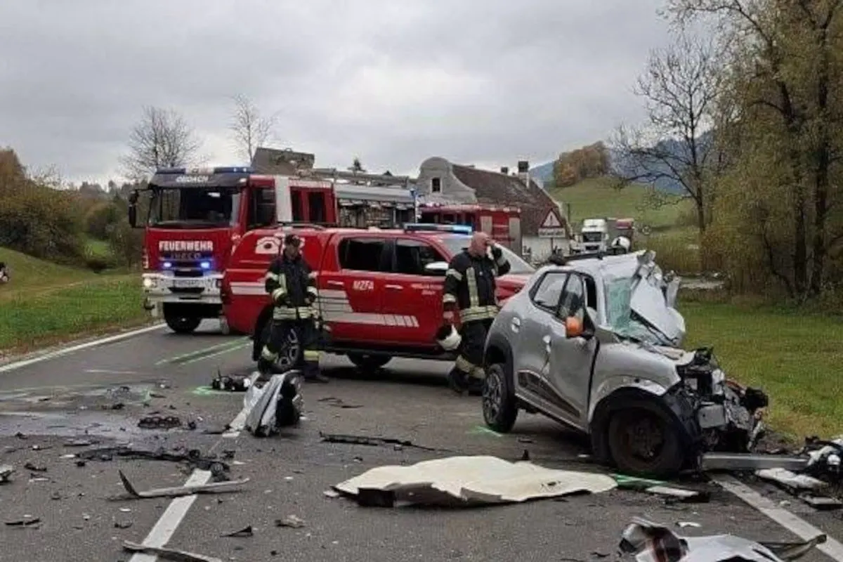 Ein stark beschädigtes silbernes Auto steht auf einer Landstraße. Zwei rote Feuerwehrfahrzeuge und Feuerwehrleute sind ebenfalls zu sehen.