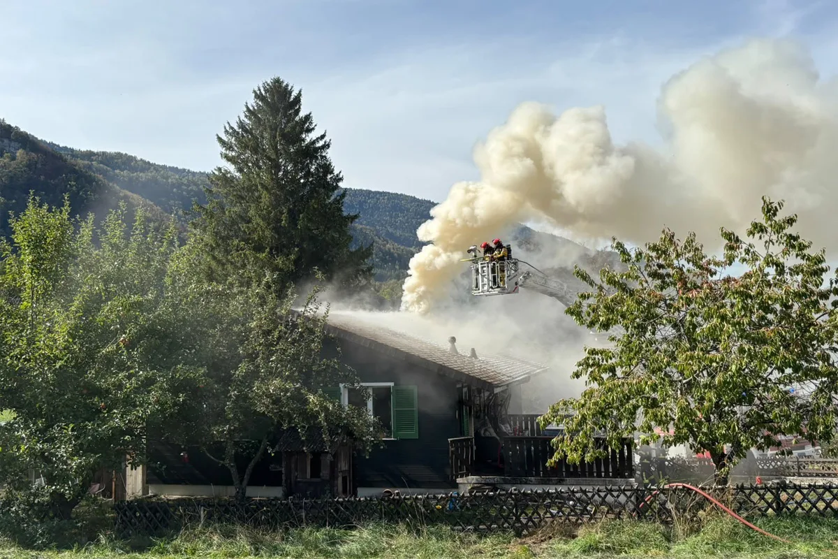 Feuerwehrleute stehen auf einer ausgefahrenen Leiter und bekämpfen starken Rauch an einem Hausdach in ländlicher Umgebung.