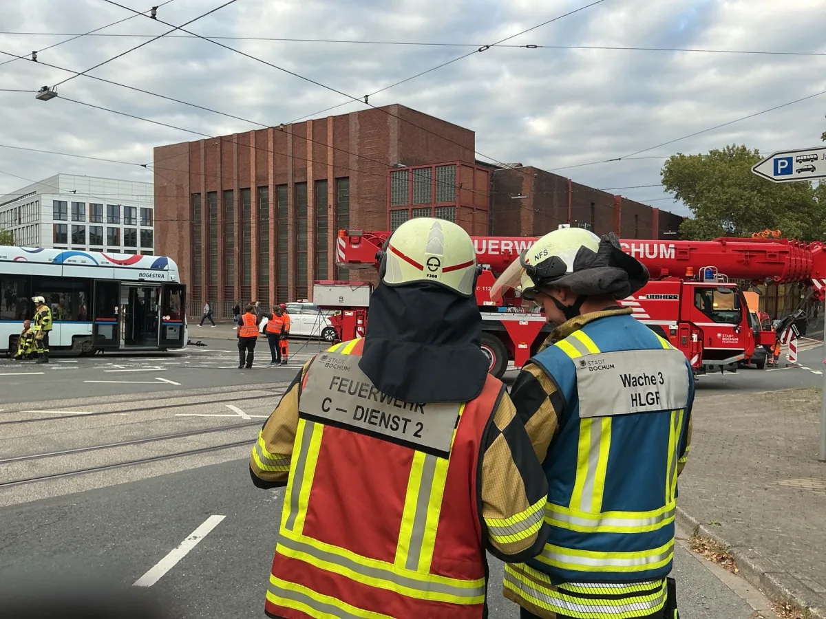 Zwei Feuerwehrmänner stehen auf einer Straße und betrachten eine Unfallstelle. Eine Straßenbahn und ein Feuerwehrfahrzeug sind im Bild sichtbar.