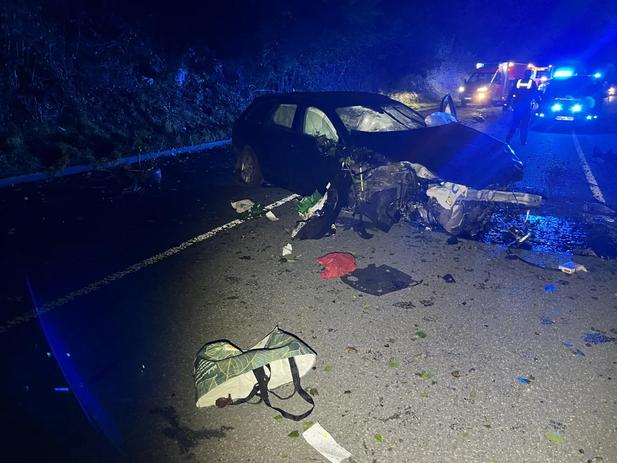 Ein stark beschädigtes Auto steht nach einem nächtlichen Unfall auf der Straße mit Trümmern umgeben. Blaulichter sind im Hintergrund sichtbar.