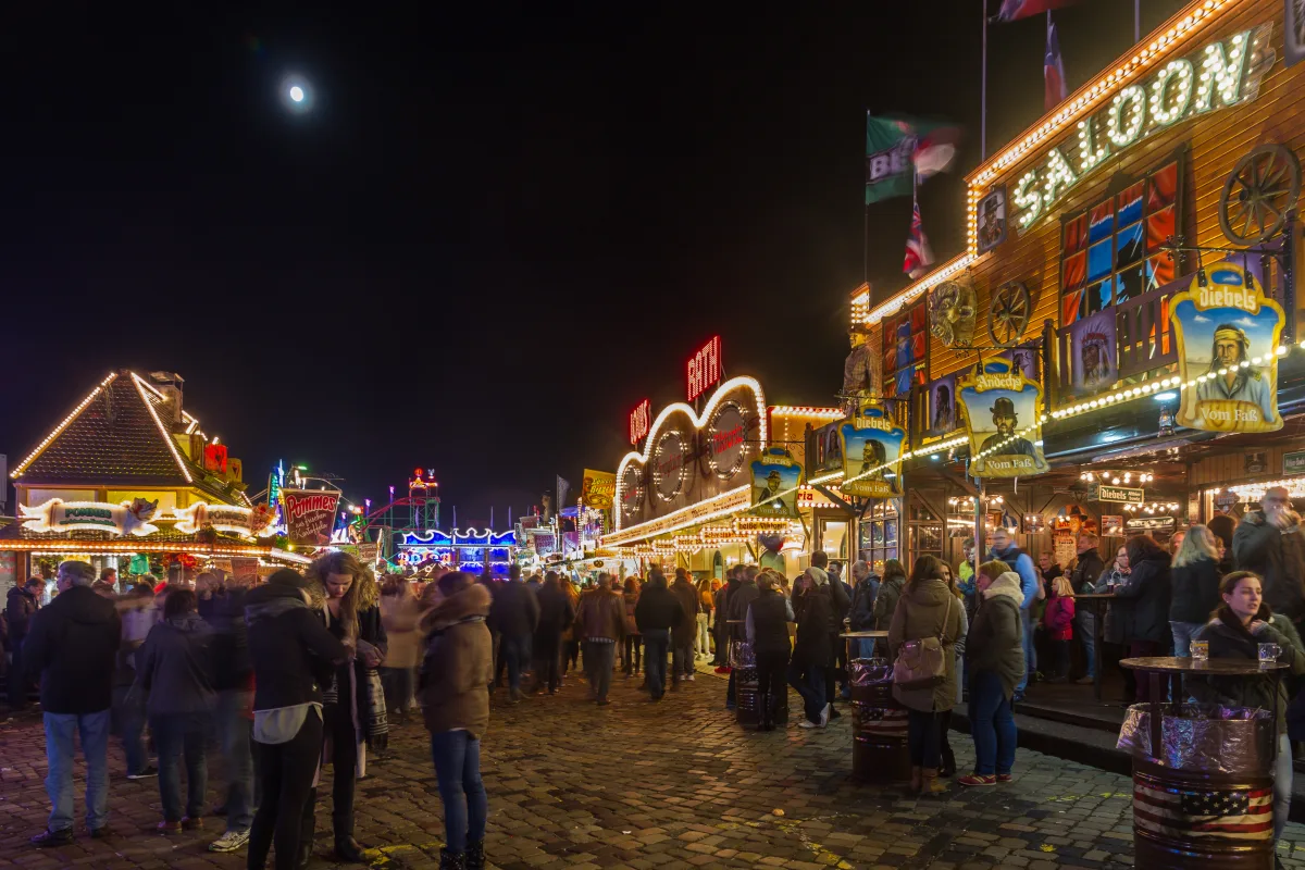 Eine Menschenmenge besucht einen hell erleuchteten Jahrmarkt bei Nacht. Verschiedene Stände und Fahrgeschäfte sind sichtbar.