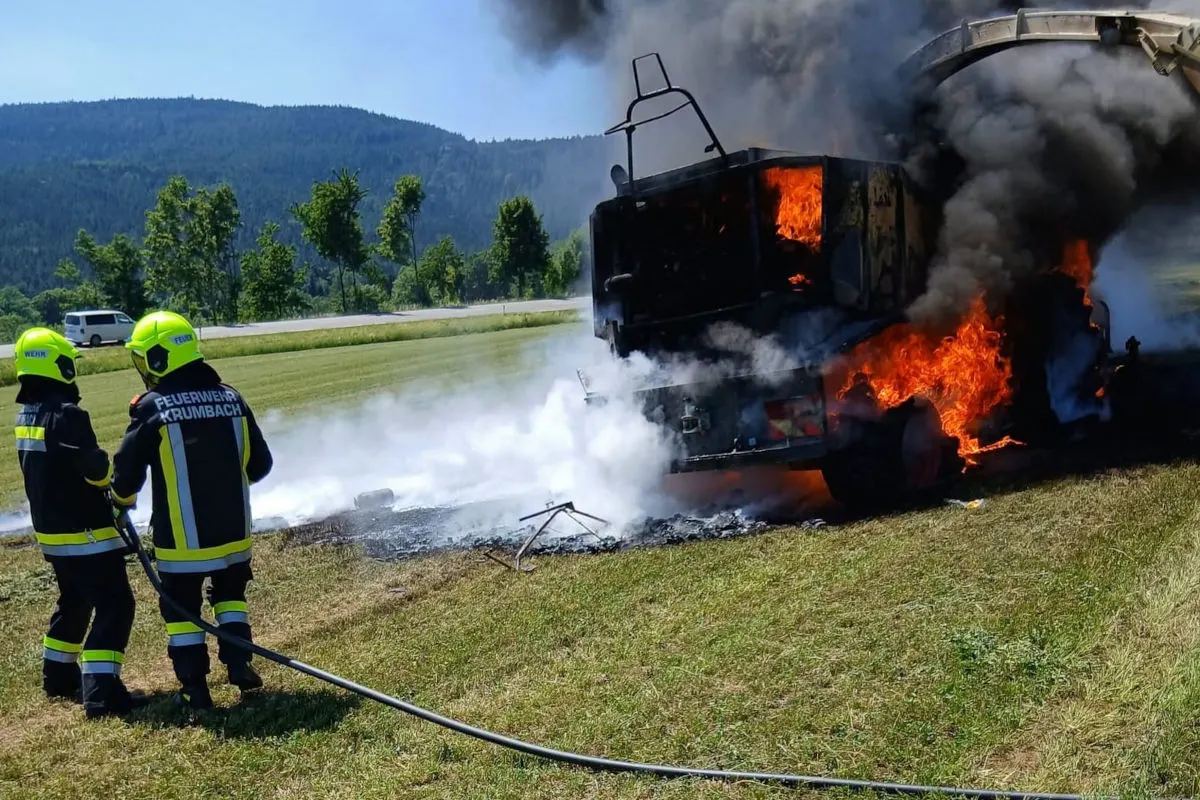 Zwei Feuerwehrleute der Feuerwehr Krumbach löschen einen stark brennenden Anhänger auf einer Wiese. Schwarzer Rauch steigt empor.