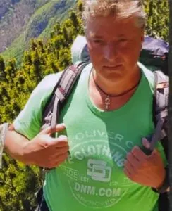 Ein Mann trägt einen Rucksack und befindet sich in einer grünen, bergigen Landschaft. Er trägt ein grünes T-Shirt und schaut direkt in die Kamera.