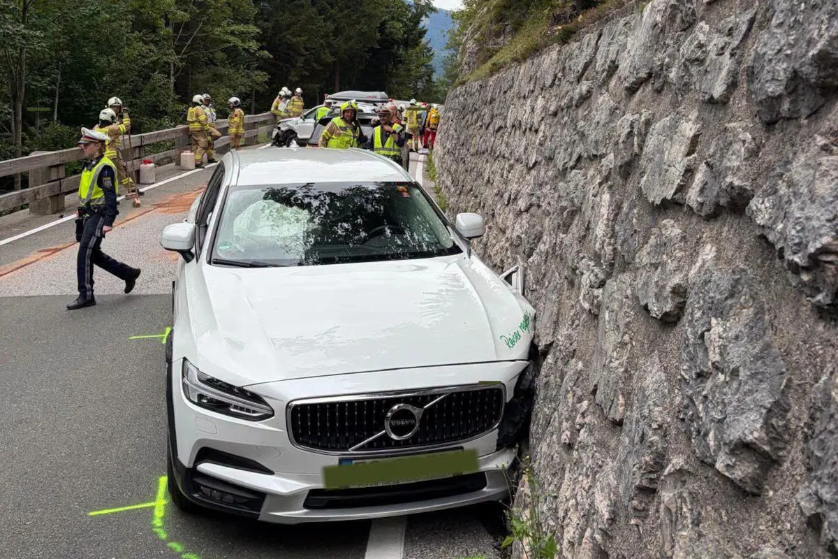 Ein weißes Volvo-Auto ist seitlich gegen eine Steinmauer geprallt. Feuerwehrleute und ein Polizist sind am Unfallort aktiv.