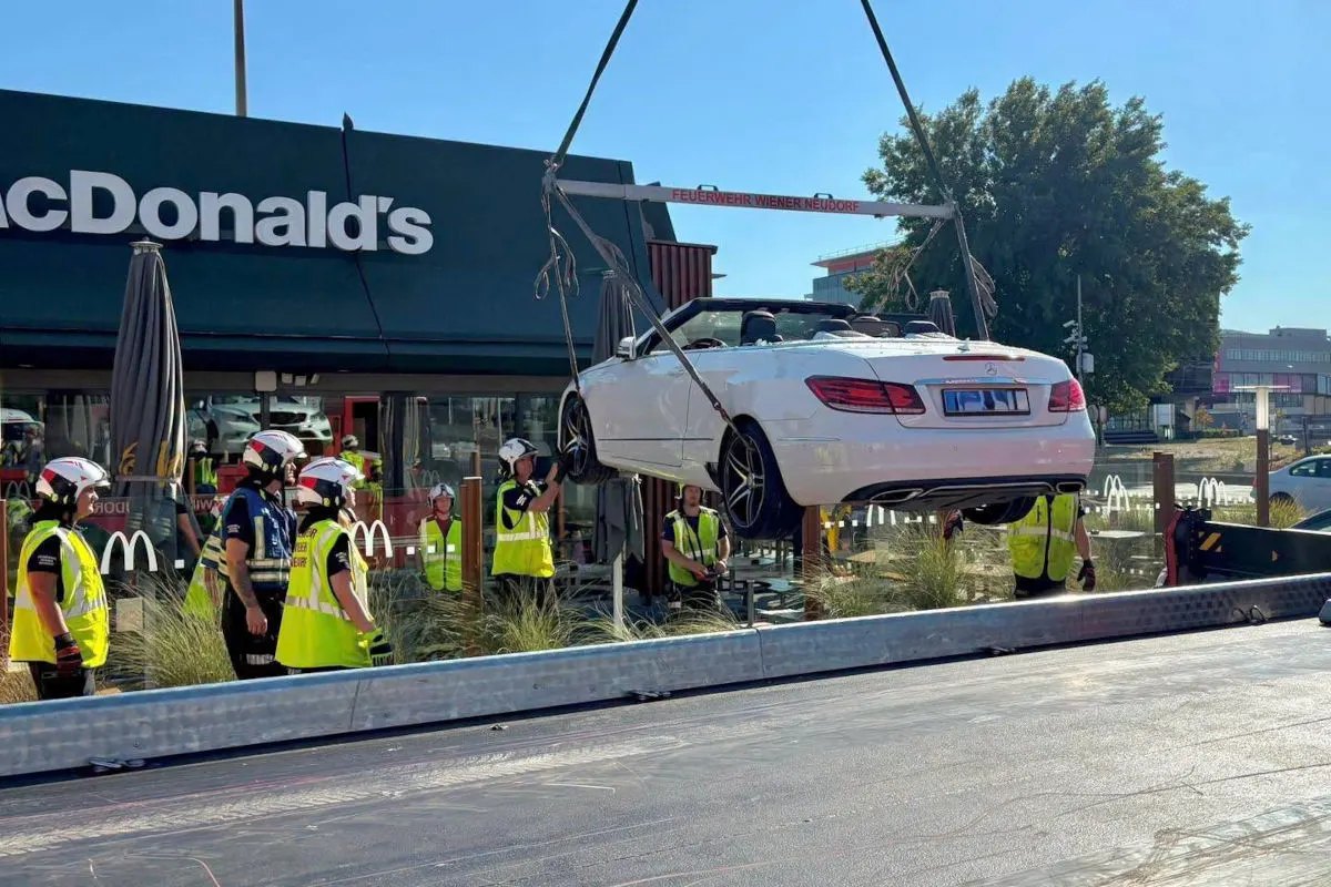 Mehrere Feuerwehrmänner heben ein weißes Cabriolet vor einem McDonald's-Restaurant mit einem Kran an. Das Fahrzeug ist beschädigt.