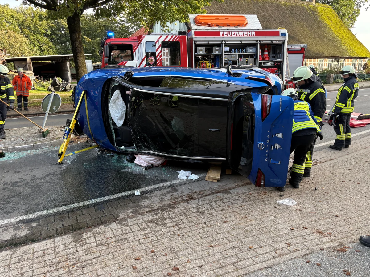 Ein Auto liegt auf der Seite auf einer Straße, umgeben von Feuerwehrleuten, die es stabilisieren und sichern. Im Hintergrund ein Feuerwehrfahrzeug.