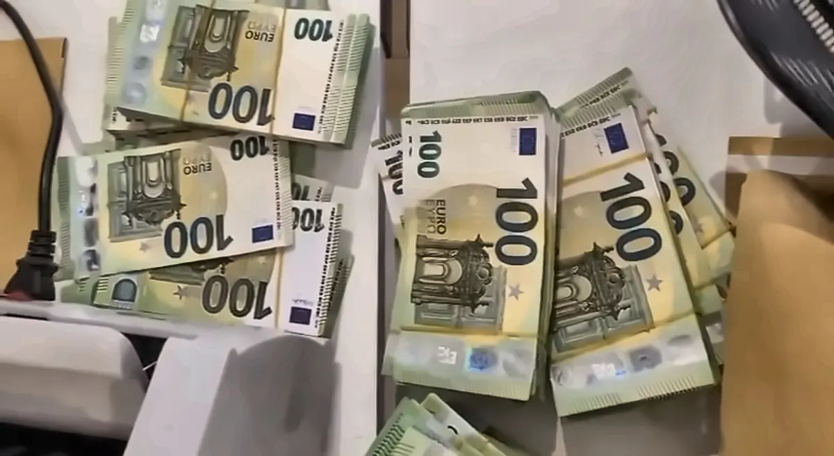 Auf einem Tisch liegen zahlreiche Stapel von 100-Euro-Banknoten. Ein Stromkabel und ein Umschlag sind ebenfalls sichtbar.