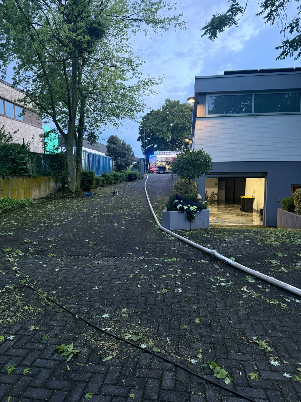 Ein Feuerwehrfahrzeug mit Blaulicht steht in einem gepflasterten Bereich vor einem Gewerbegebäude. Der Boden ist mit Blättern bedeckt, die durch den Wind verteilt wurden.