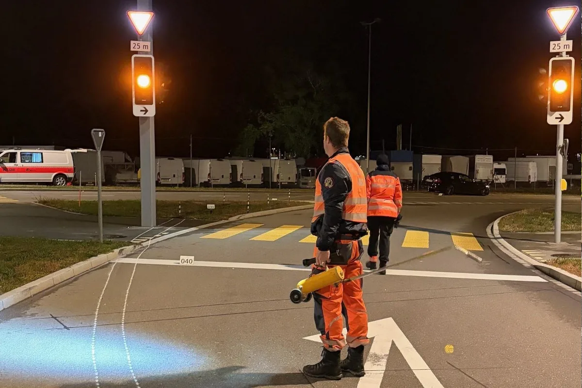 Zwei Männer in orangen reflektierenden Westen stehen auf einer nächtlichen Straßenkreuzung. Einer hält ein Megaphon.