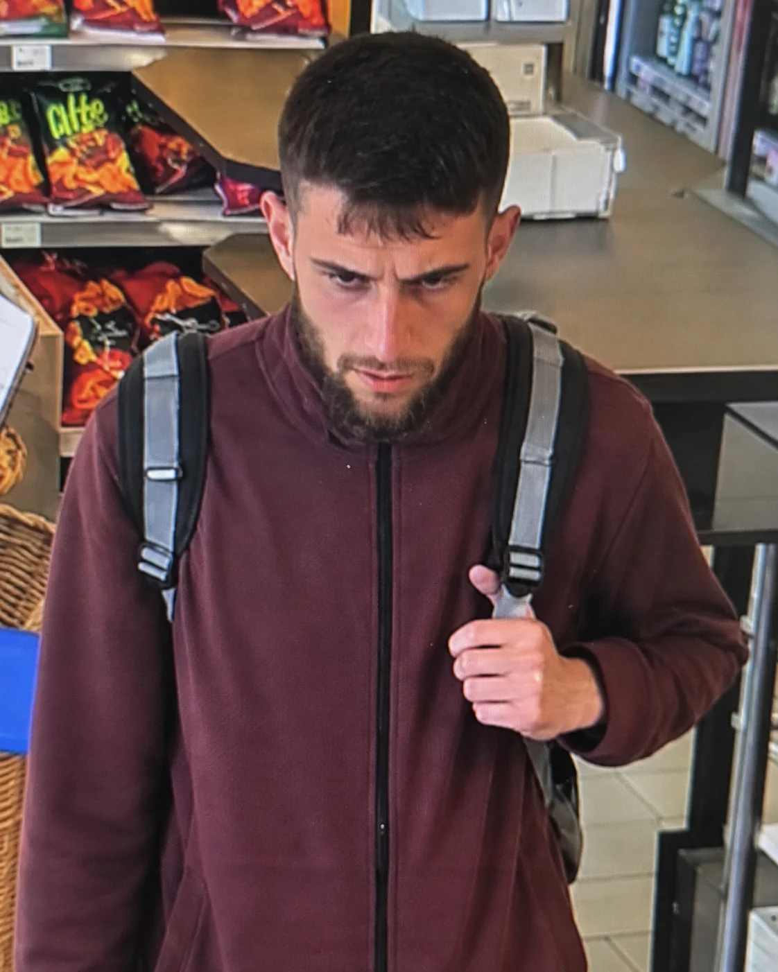 Ein Mann mit Bart und Rucksack steht in einem Geschäft vor einem Regal mit Snacks. Er schaut konzentriert nach unten.