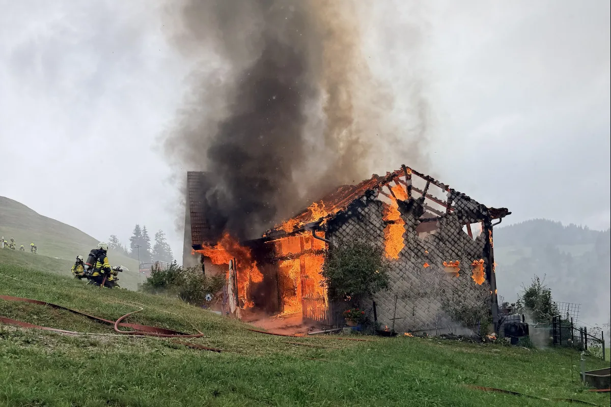 Ein größeres Holzhaus steht in Flammen, Flammen und Rauch steigen auf. Feuerwehrleute befinden sich am Hang und löschen den Brand.