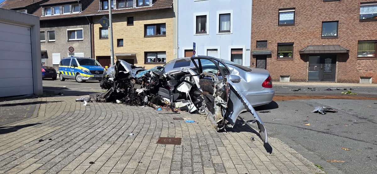 Ein stark beschädigtes Auto steht auf einer Straßenkreuzung. Im Hintergrund ist ein deutscher Polizei-Van erkennbar.