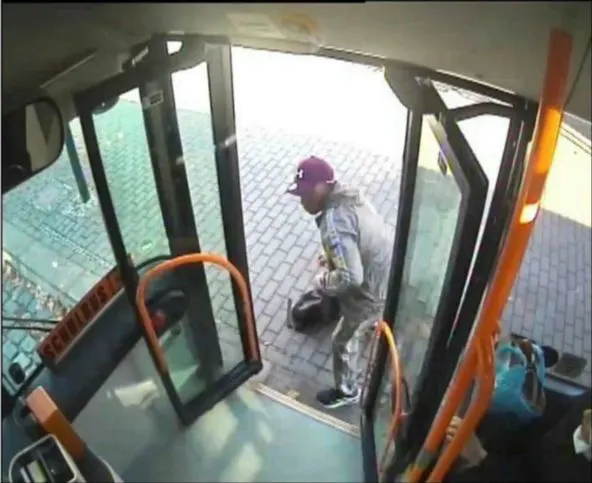 Ein Mann mit einer Baseballkappe und Trainingsanzug steigt in einen Bus ein und trägt eine Tasche. Die Szene wird von einer Überwachungskamera aufgenommen.