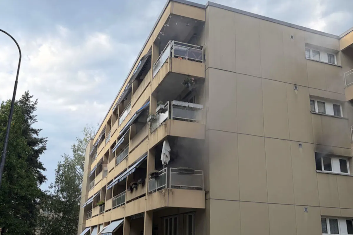 Ein mehrstöckiges Wohnhaus mit Rauch, der aus einem der Balkone aufsteigt. Der Himmel ist bewölkt und Bäume sind neben dem Gebäude sichtbar.