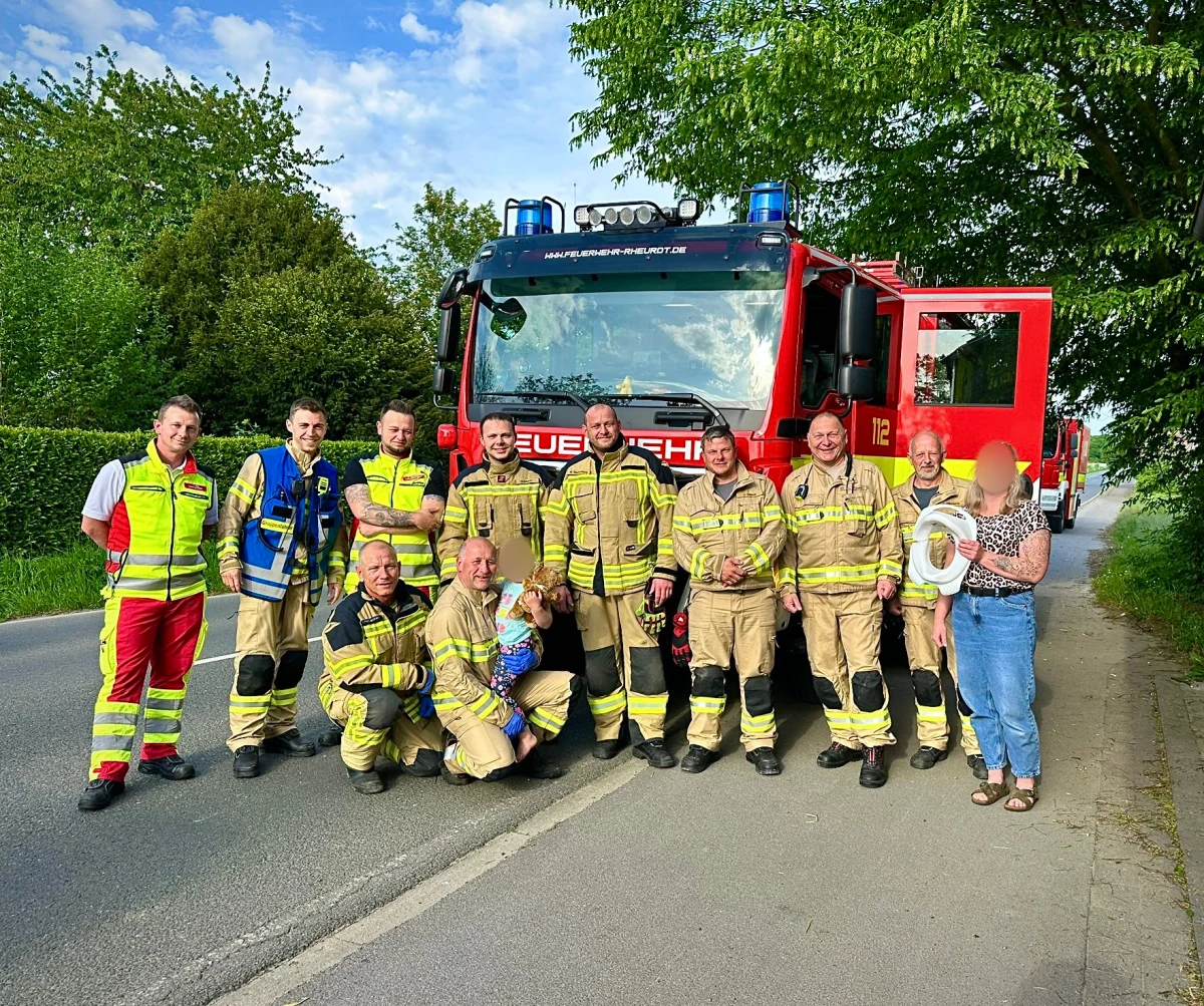 Eine Gruppe von Feuerwehrleuten in Einsatzkleidung steht auf einer Straße vor einem roten Feuerwehrfahrzeug mit Aufschrift. Sie lächeln und posieren für das Foto.