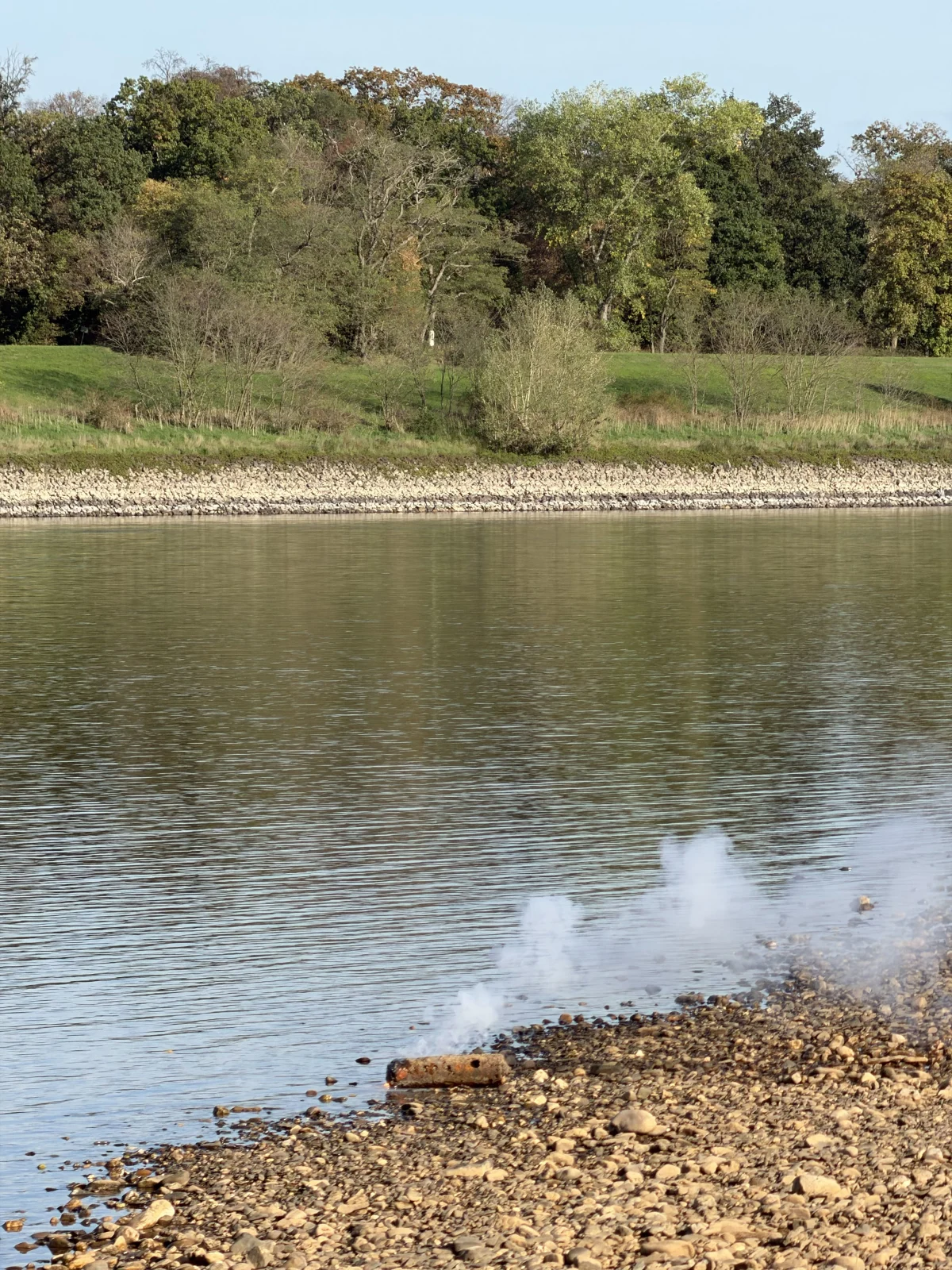 Ein rauchendes, zylinderförmiges Objekt liegt nahe am Ufer eines Flusses, umgeben von Kieselsteinen und Wasser. Im Hintergrund ist eine bewaldete Uferlandschaft zu sehen.