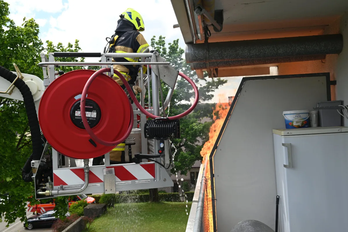 Ein Feuerwehrmann in Schutzkleidung auf einem Leiterfahrzeug löscht ein Feuer auf einem Balkon. Es sind Flammen sichtbar sowie ein Kühlschrank und andere Haushaltsgegenstände.