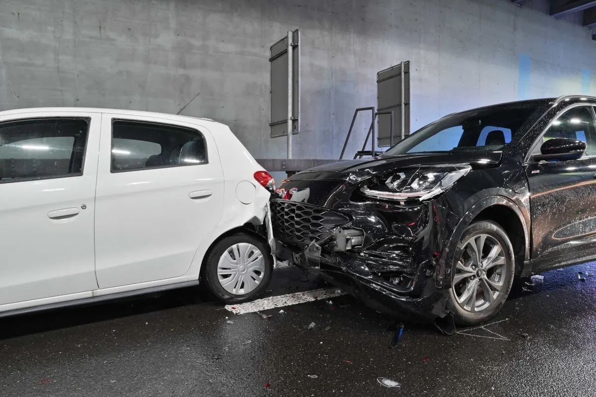 Ein weißer Kleinwagen und ein schwarzes SUV sind in einem Tunnel frontal kollidiert. Beide Fahrzeuge haben sichtbare Schäden an der Front.