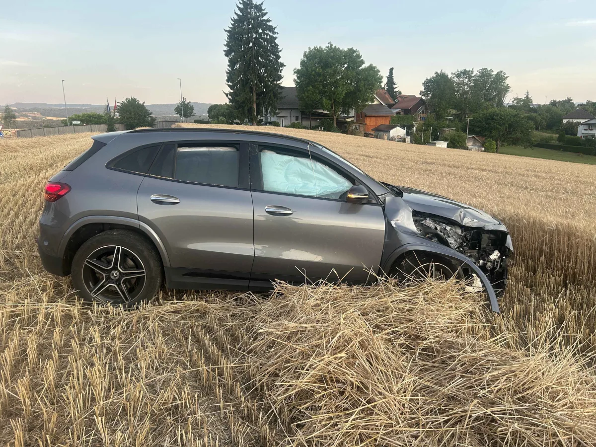 Ein graues Fahrzeug mit stark beschädigter Front steht in einem abgeernteten Feld. Im Hintergrund sind Bäume und eine Wohnsiedlung sichtbar.