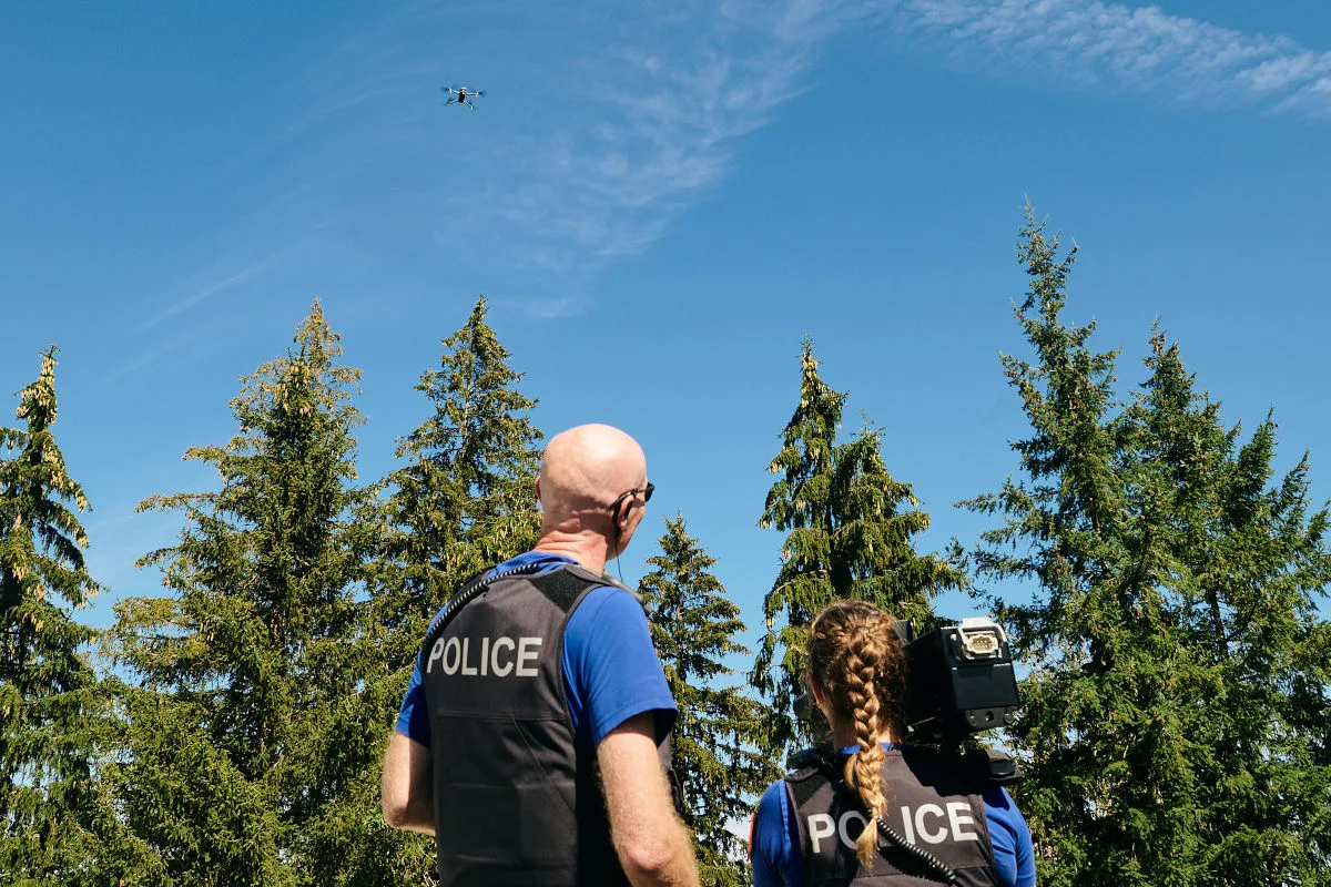 Zwei Polizisten in Uniform stehen im Wald und schauen eine fliegende Drohne an. Der Himmel ist klar und blau, umgeben von hohen Nadelbäumen.