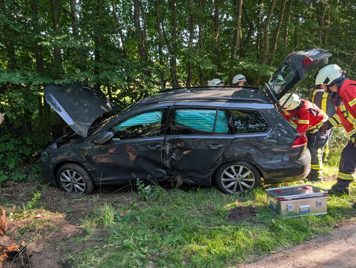 Ein beschädigter dunkler Kombi steht an einem Waldstück, mehrere Feuerwehrleute in Einsatzkleidung sind vor Ort.