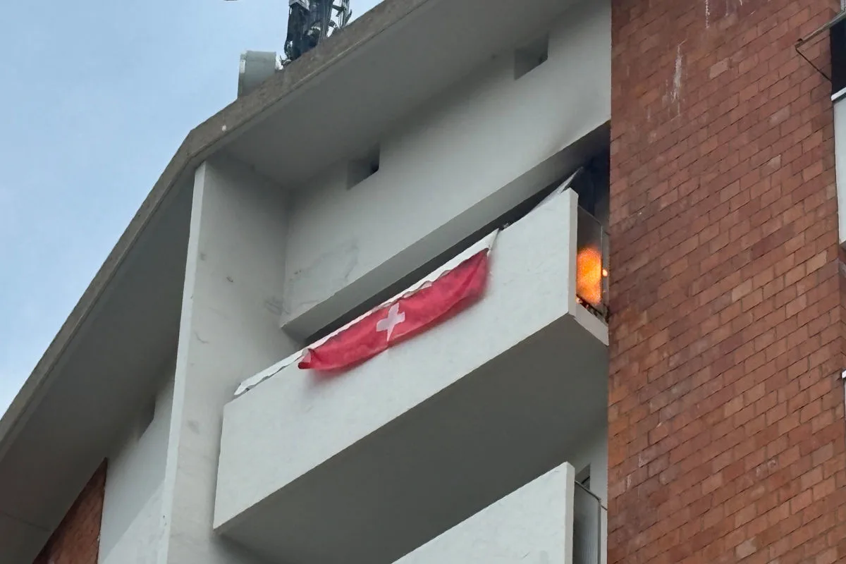 Ein Balkon eines Wohnhauses brennt, eine Schweizer Flagge hängt darüber. Der Brand scheint aus einem oberen Geschoss zu stammen.