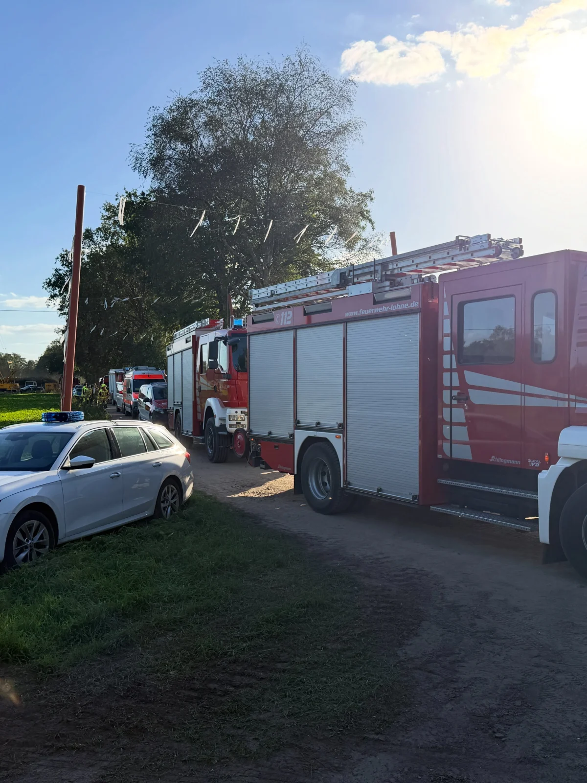 Zwei rote Feuerwehrwagen und ein weißes Polizeiauto parken auf einem Feldweg neben grünem Gras unter blauem Himmel.