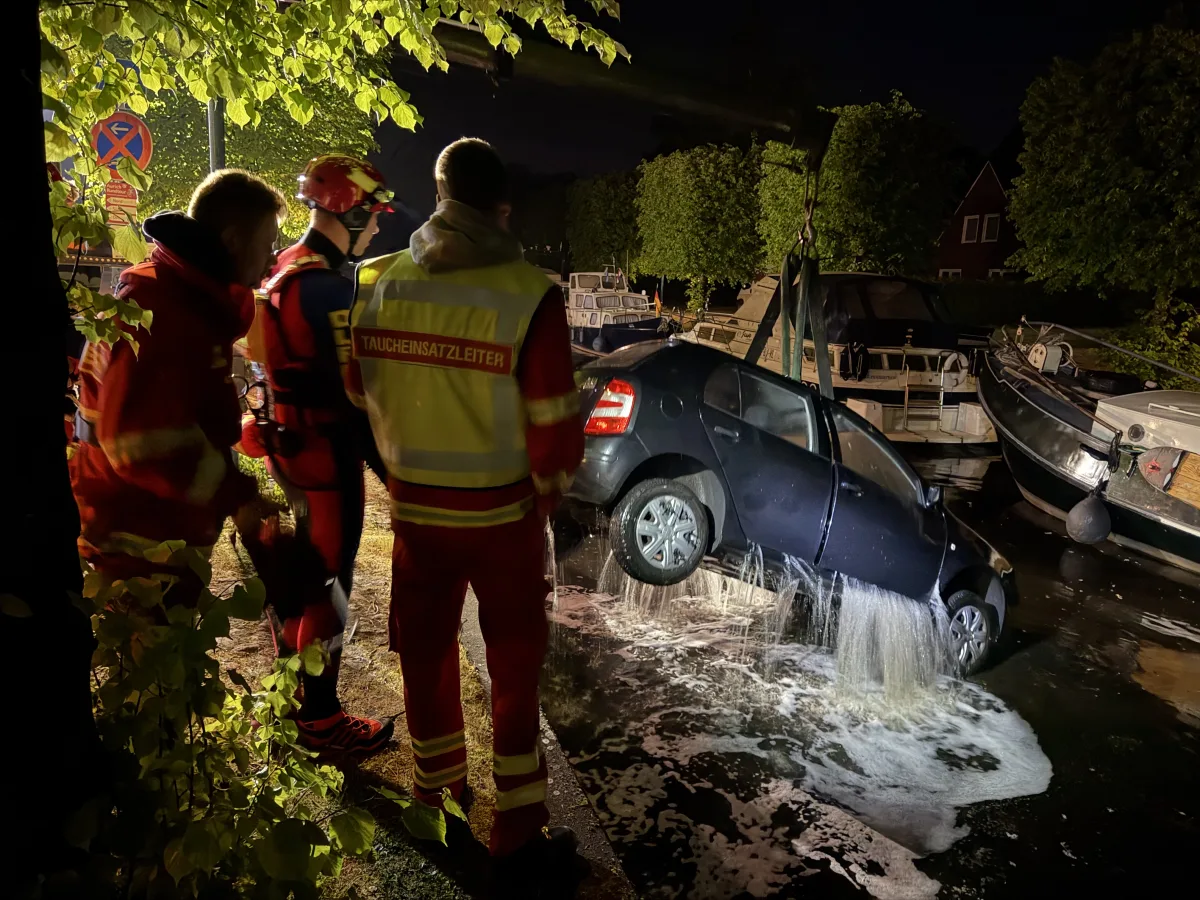 Drei Einsatzkräfte stehen an einem Kanal, während ein Auto aus dem Wasser gehoben wird. Die Szene ist nachts beleuchtet.