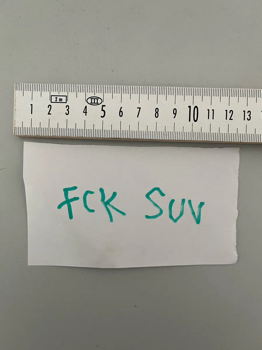 Ein Zettel mit der handschriftlichen Aufschrift 'FCK SUV' liegt neben einem Lineal auf einer grauen Oberfläche.
