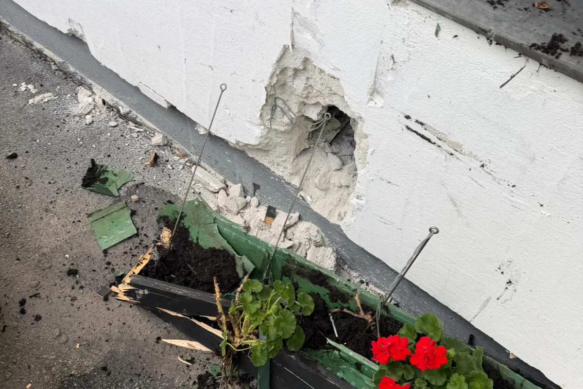 Eine weiße Wand zeigt einen großen Schaden, daneben liegt ein beschädigter Blumenkasten mit roten Blumen auf dem Boden.