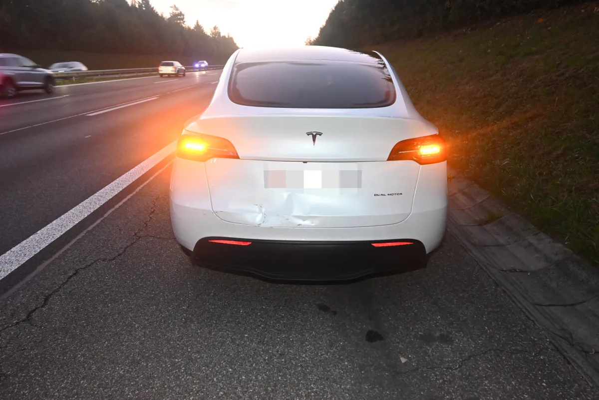 Ein weißer Tesla mit sichtbarem Heckschaden steht am Standstreifen einer Autobahn. Im Hintergrund sind vorbeifahrende Autos zu sehen, der Himmel deutet Dämmerung an.