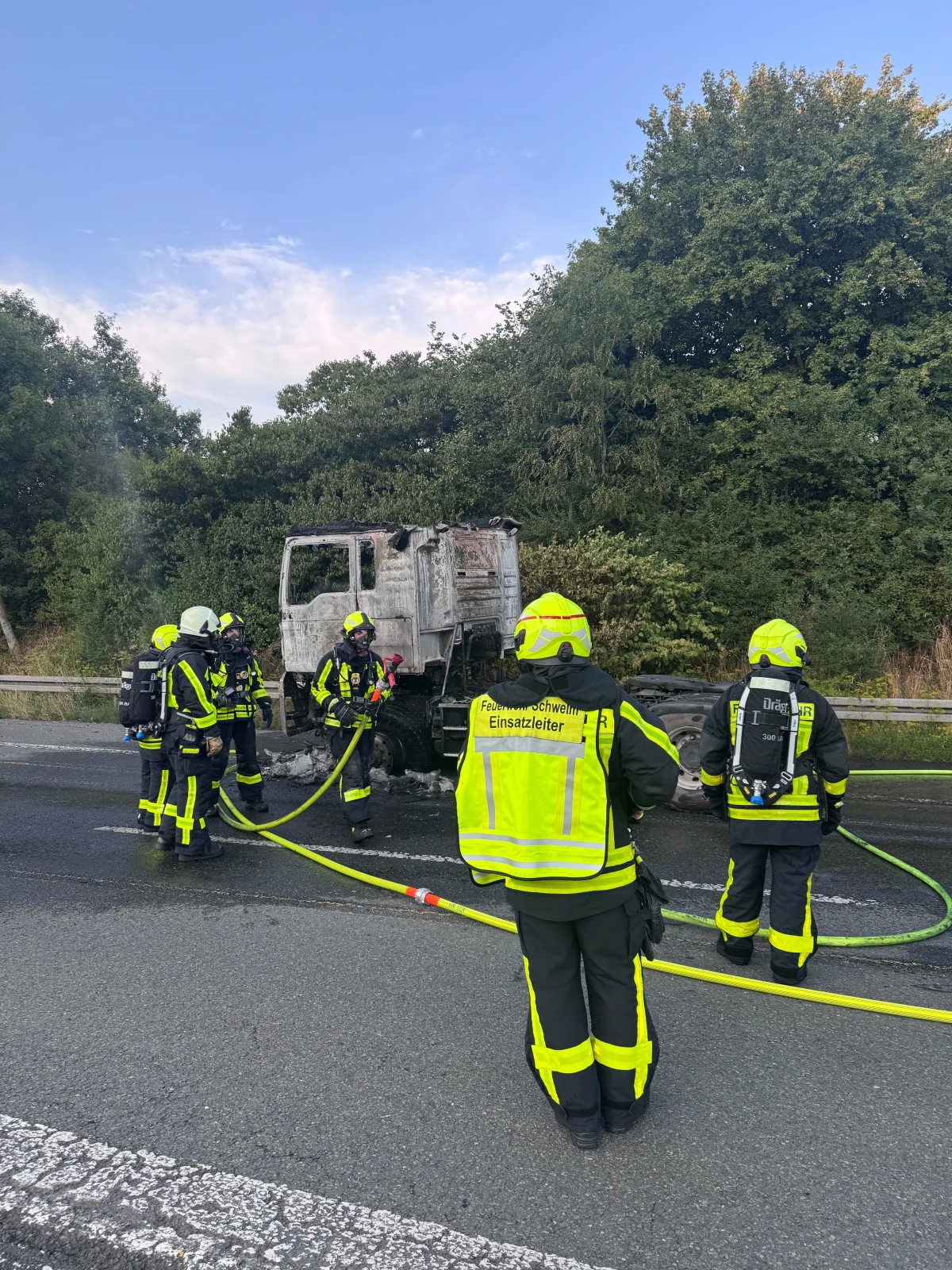 Mehrere Feuerwehrleute löschen mit Schläuchen einen ausgebrannten Lkw auf einer Landstraße. Dichter Wald im Hintergrund.