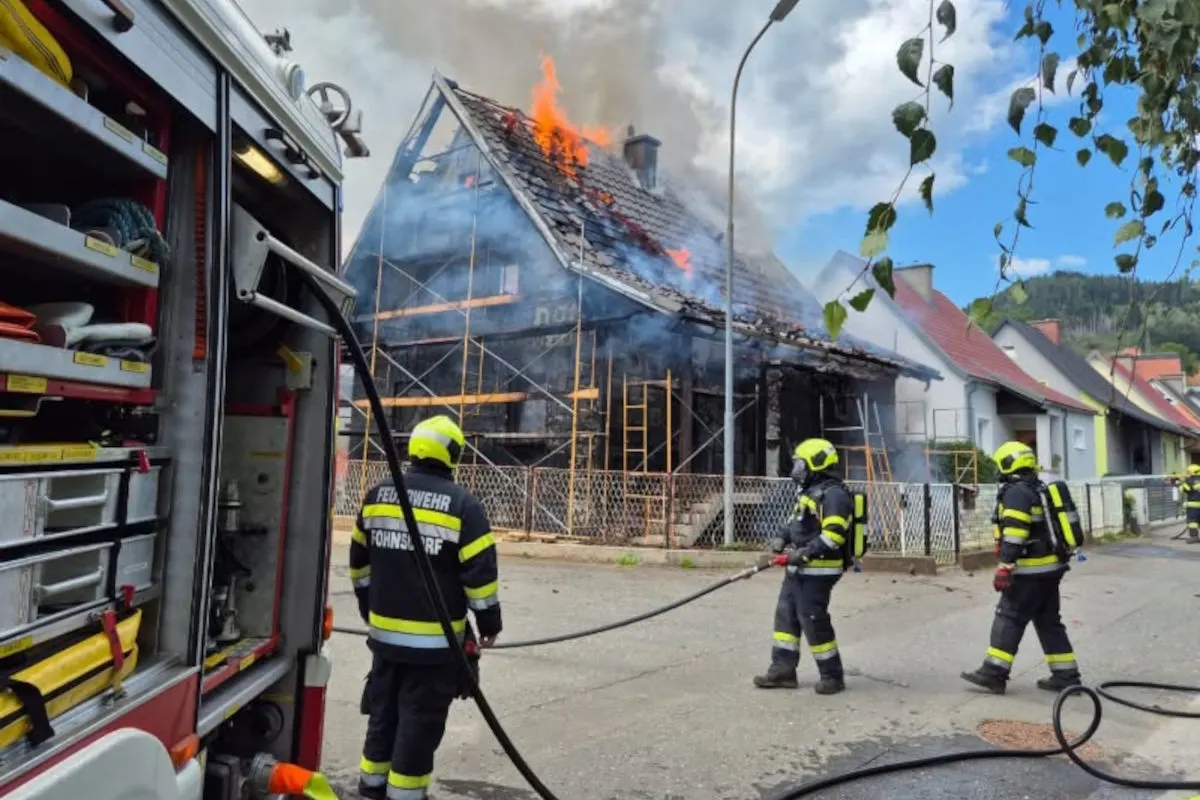 Mehrere Feuerwehrleute in Schutzkleidung bekämpfen einen Dachbrand eines Hauses mit offenen Flammen und Rauchentwicklung.