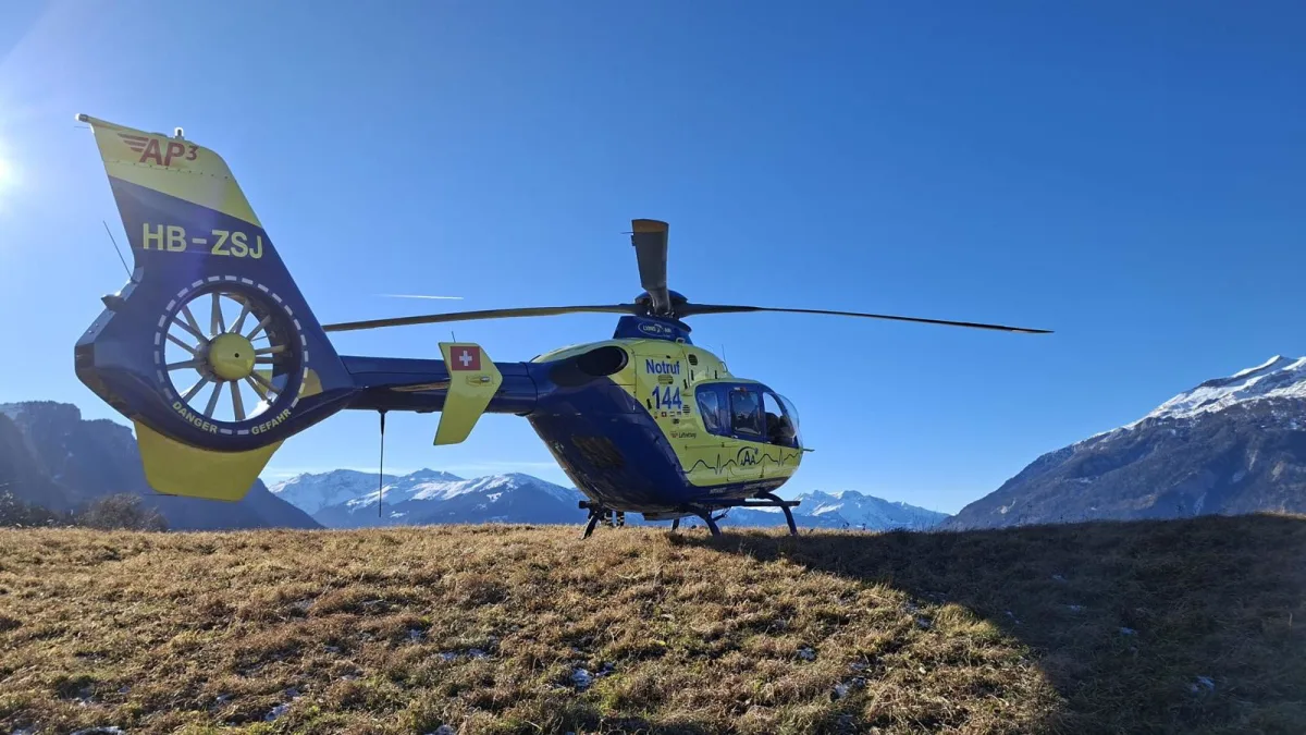 Ein gelber und blauer Rettungshubschrauber steht auf einem Bergplateau. Im Hintergrund sieht man schneebedeckte Berge unter klarem Himmel.
