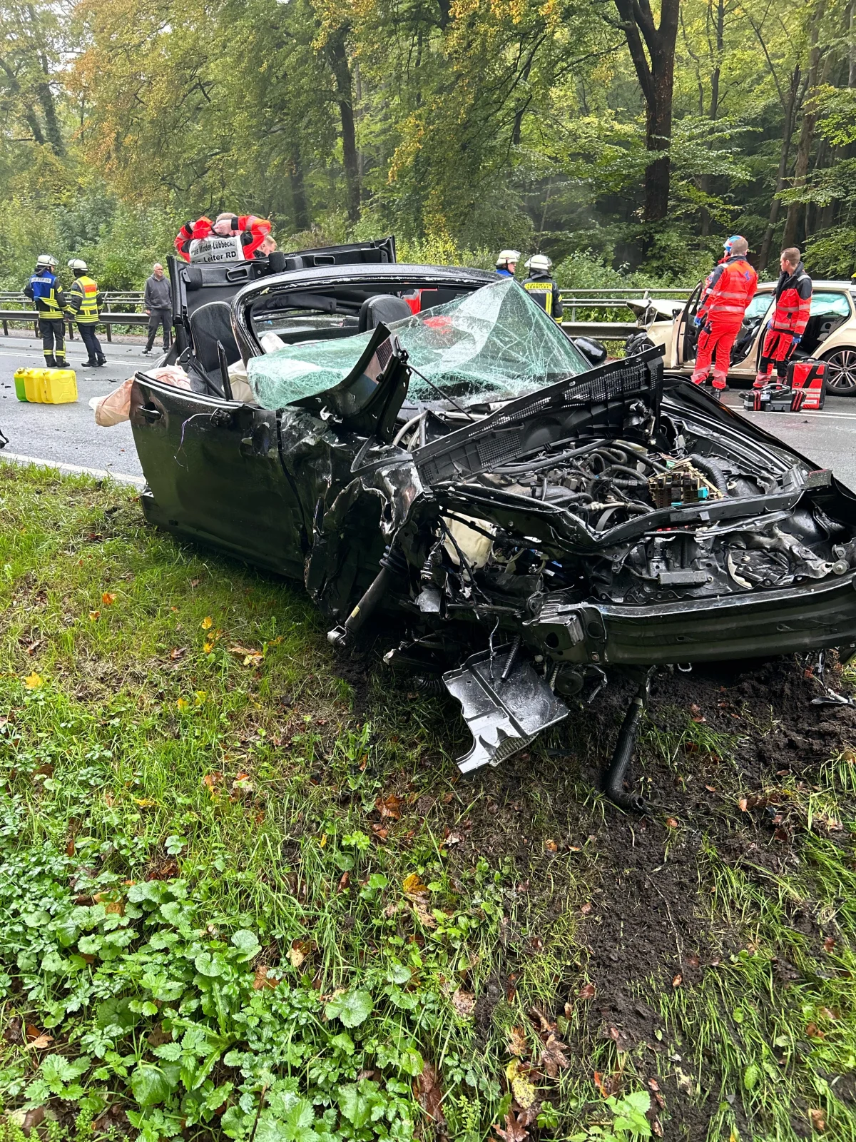 Ein stark beschädigtes schwarzes Fahrzeug steht am Straßenrand, von Rettungskräften umgeben. Im Hintergrund sind Bäume sichtbar.