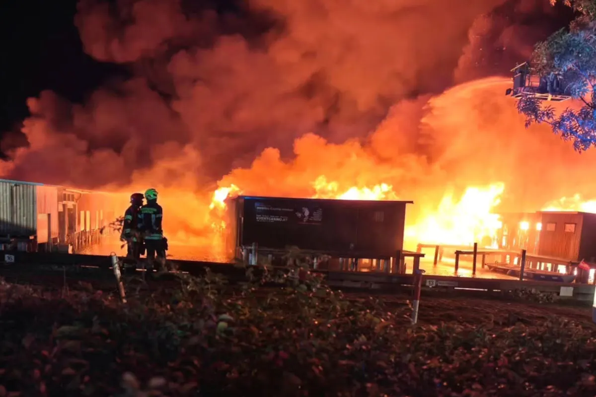 Im Vordergrund stehen zwei Feuerwehrleute, die auf ein intensives Feuer blicken, das ein Gebäude in der Nacht einhüllt.
