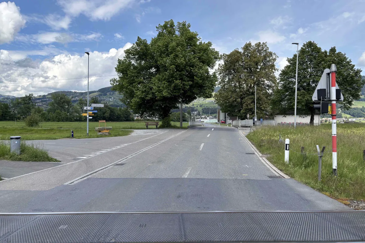 Eine Landstraße mit einem Bahnübergang im Vordergrund. Bäume flankieren die Straße, und es ist ein klarer, blauer Himmel sichtbar.