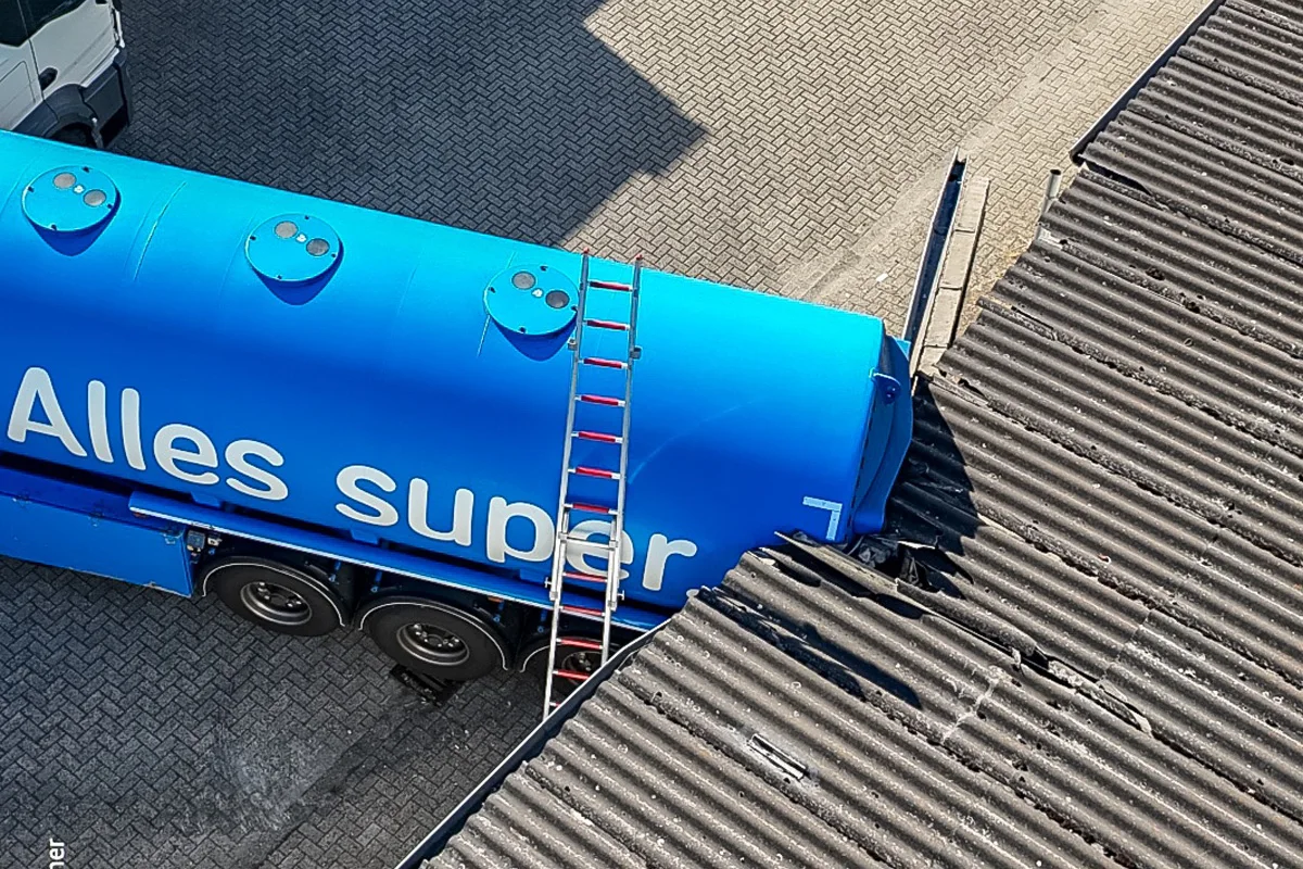 Ein blauer Tanklaster mit der Aufschrift 'Alles super' ist gegen ein Wellblechdach gefahren, wobei es beschädigt wurde. Der Laster steht nahe einer gepflasterten Fläche, und eine Aluminiumleiter ist angelehnt.