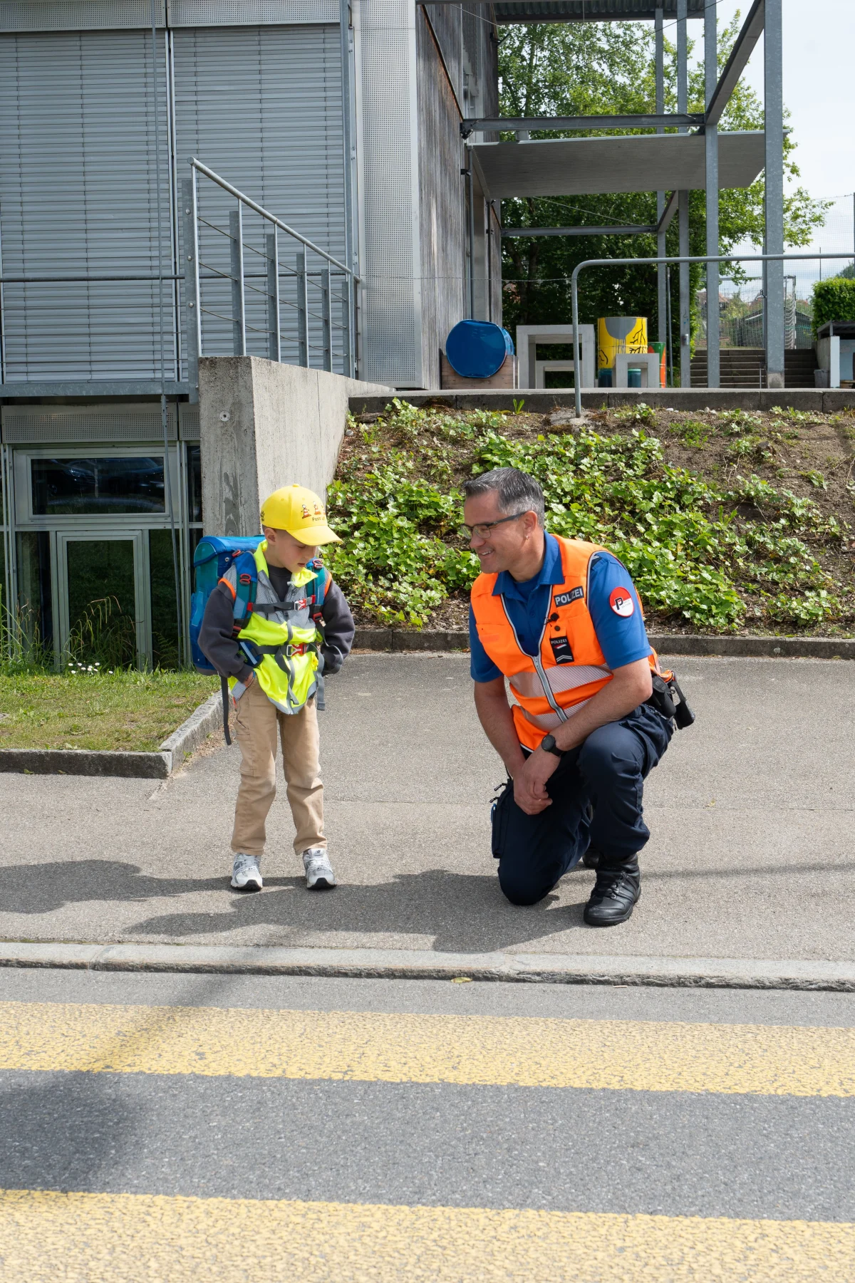 Ein Polizist in einer orangenen Warnweste kniet und spricht mit einem Schulkind, das einen Rucksack trägt.