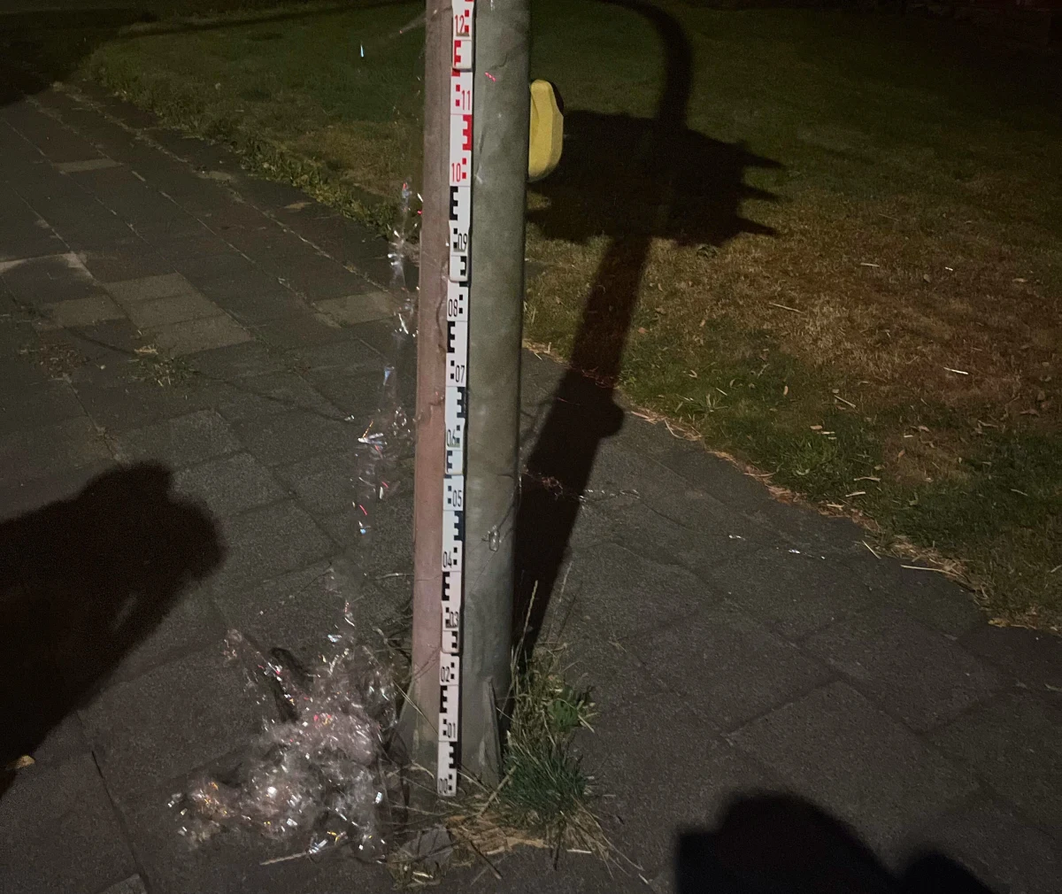 Ein Mast steht auf einem Gehweg, umgeben von Plastikfolie unter einer polizeilichen Messlatte. Die Szene ist bei Nacht aufgenommen.