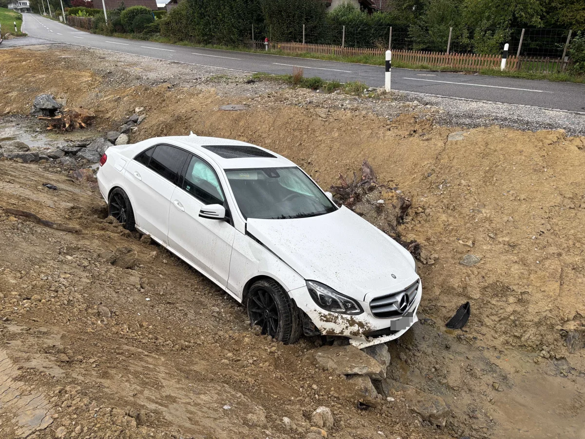 Ein weißes Auto der Marke Mercedes ist in einer Baugrube neben einer Landstraße festgefahren. Das Fahrzeug hat Matsch und Schmutz auf der Karosserie.